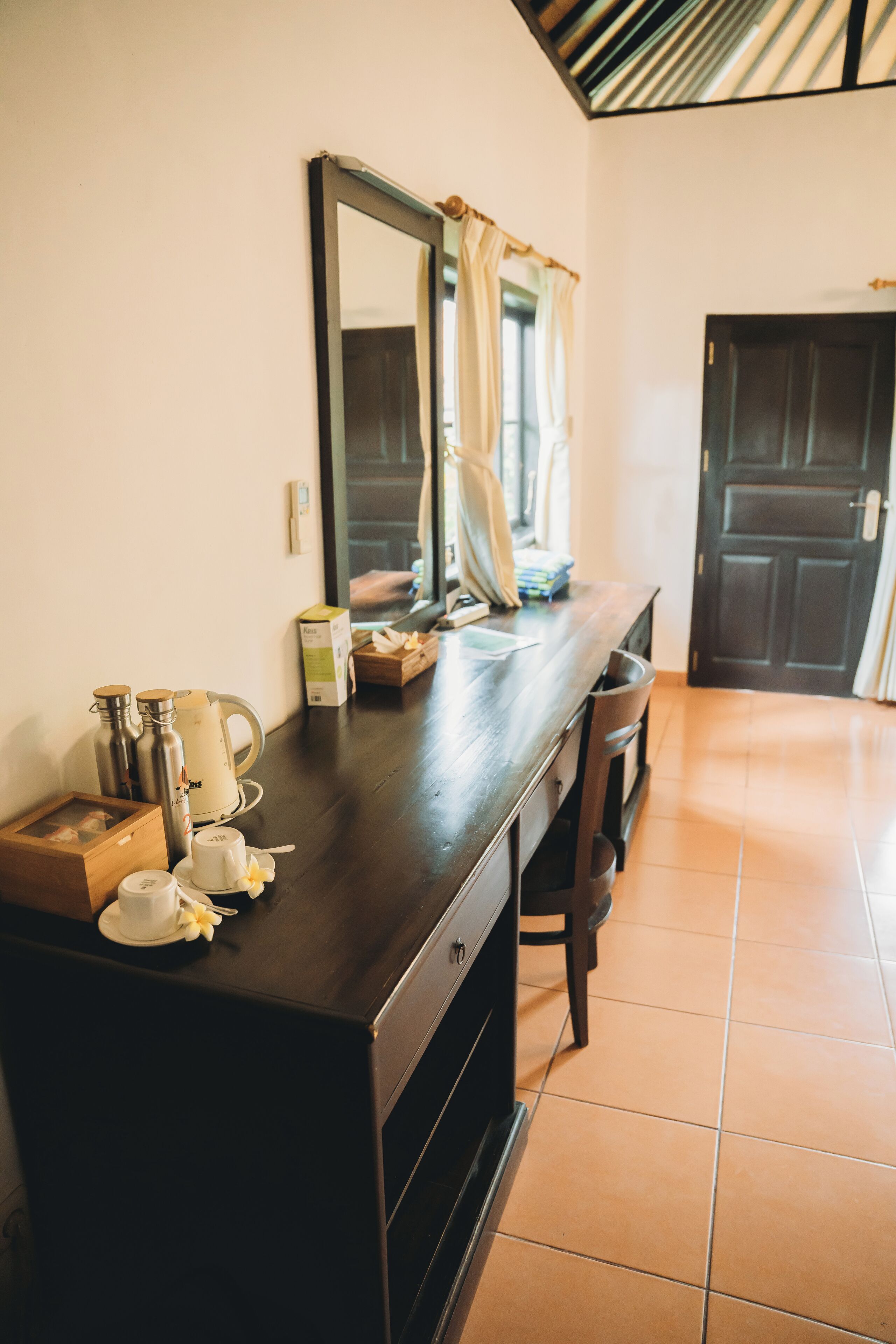 A wooden dresser with a mirror, a kettle, and a cup in a room with tiled flooring and a dark door.