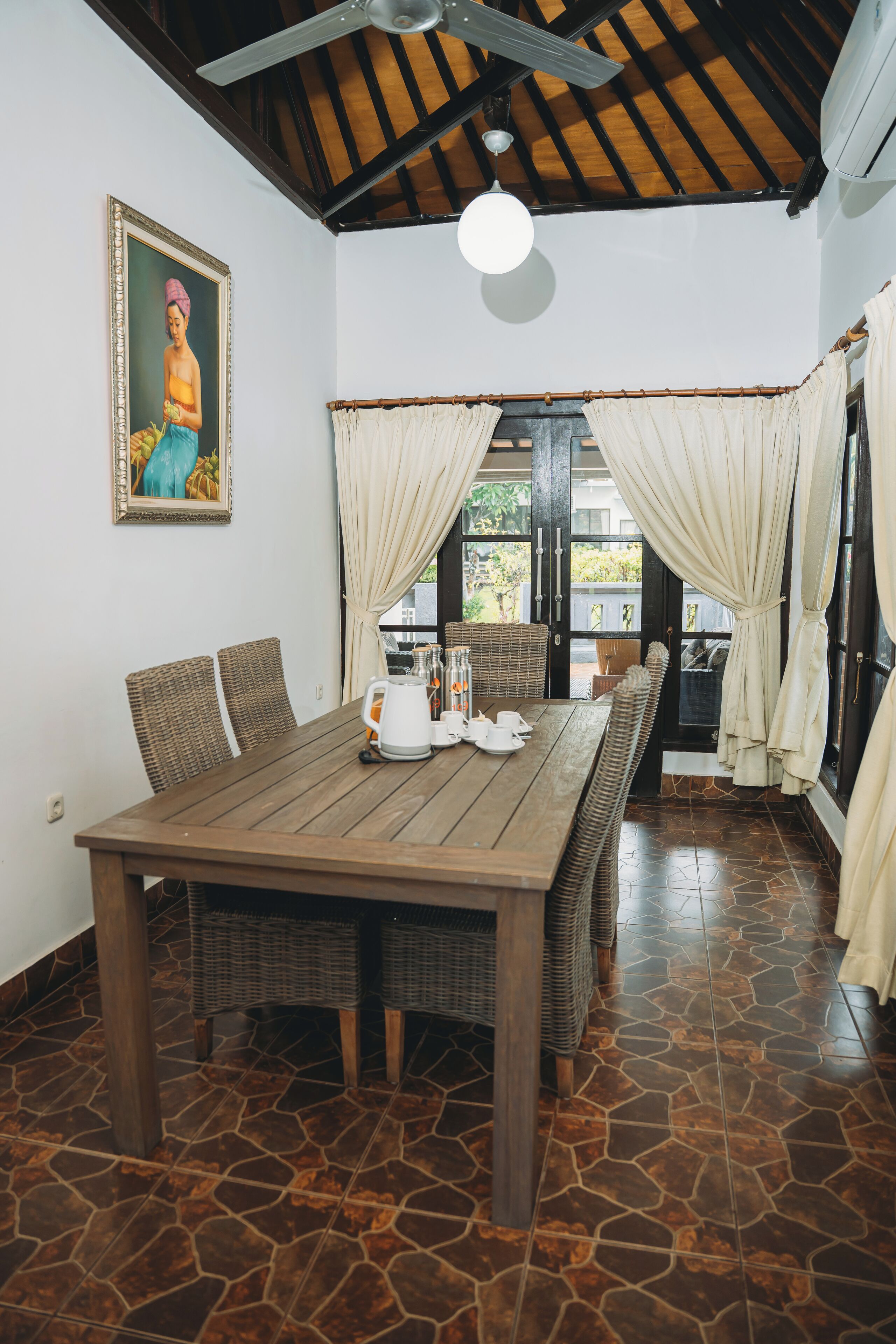 A dining area with a wooden table, wicker chairs, a painting on the wall, and a view through the door.