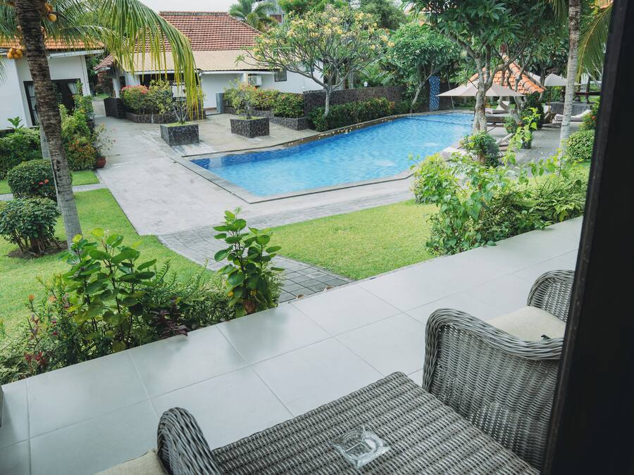 A swimming pool surrounded by lush greenery, with a patio area featuring wicker furniture and a clear glass bowl.