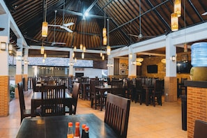 An indoor dining area with wooden tables and chairs, hanging pendant lights, and a high ceiling with exposed beams. - Liberty Dive Resort (Tulamben)