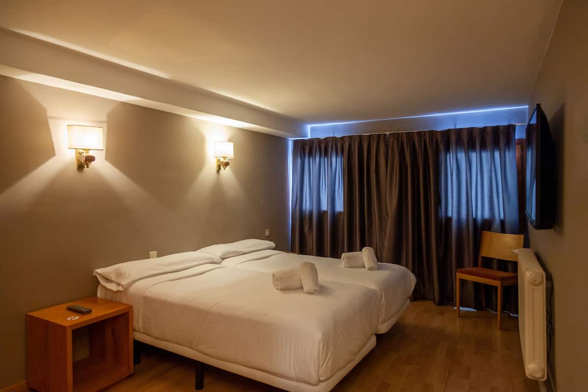 Une chambre d’hôtel avec deux lits, une table de chevet en bois, un téléviseur fixé au mur et une fenêtre avec des rideaux.