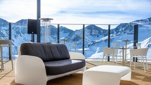 Ein stilvoller Lounge-Bereich mit einer Couch, Hocker, einem Tisch und Stühlen mit Blick auf eine verschneite Berglandschaft.