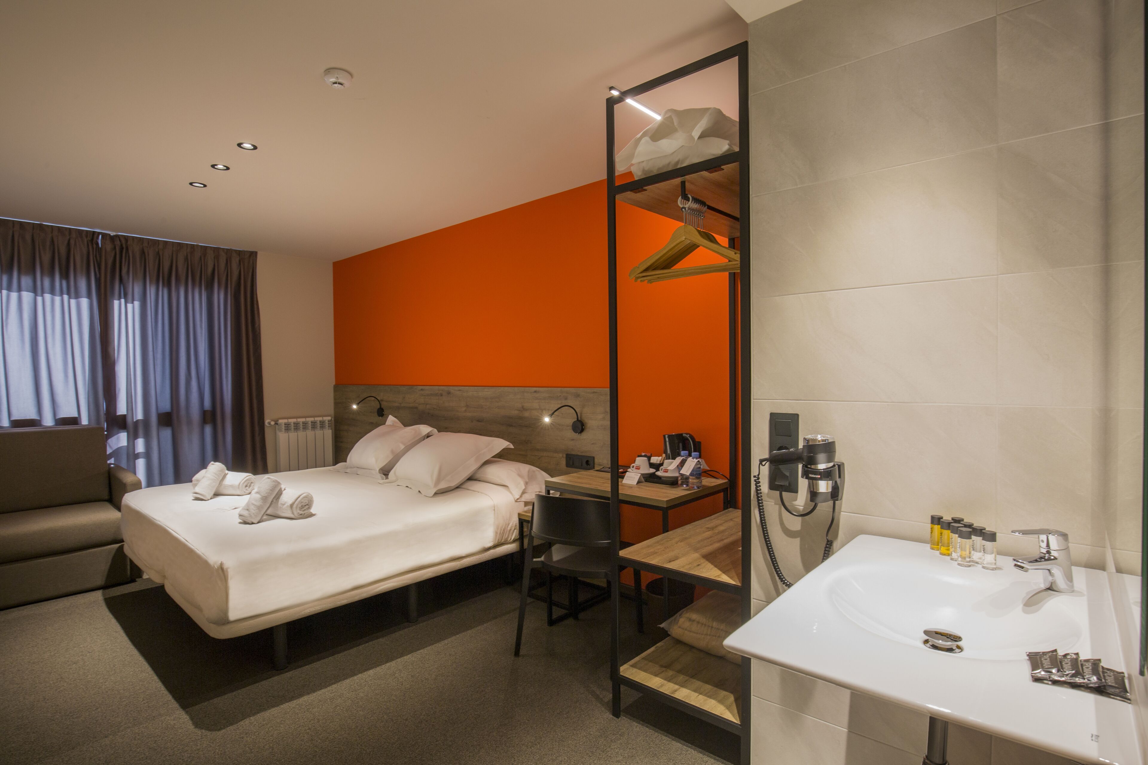 A hotel room with two beds, a desk, a chair, a bathroom with a sink, and a mirror.