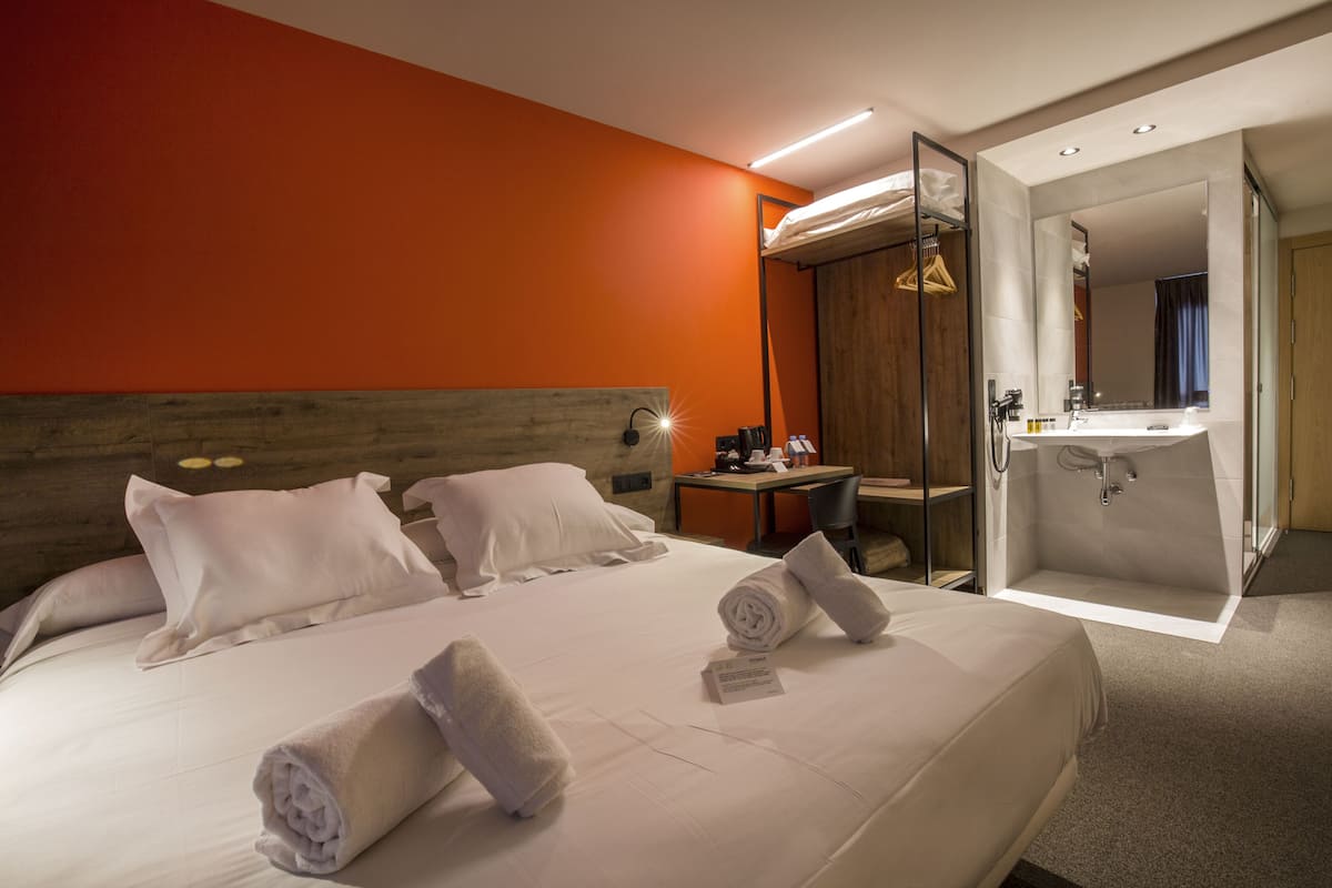 Une chambre d’hôtel avec un grand lit, une tête de lit en bois, un bureau avec une chaise, une salle de bain avec un lavabo et un miroir, et une vue sur la ville.