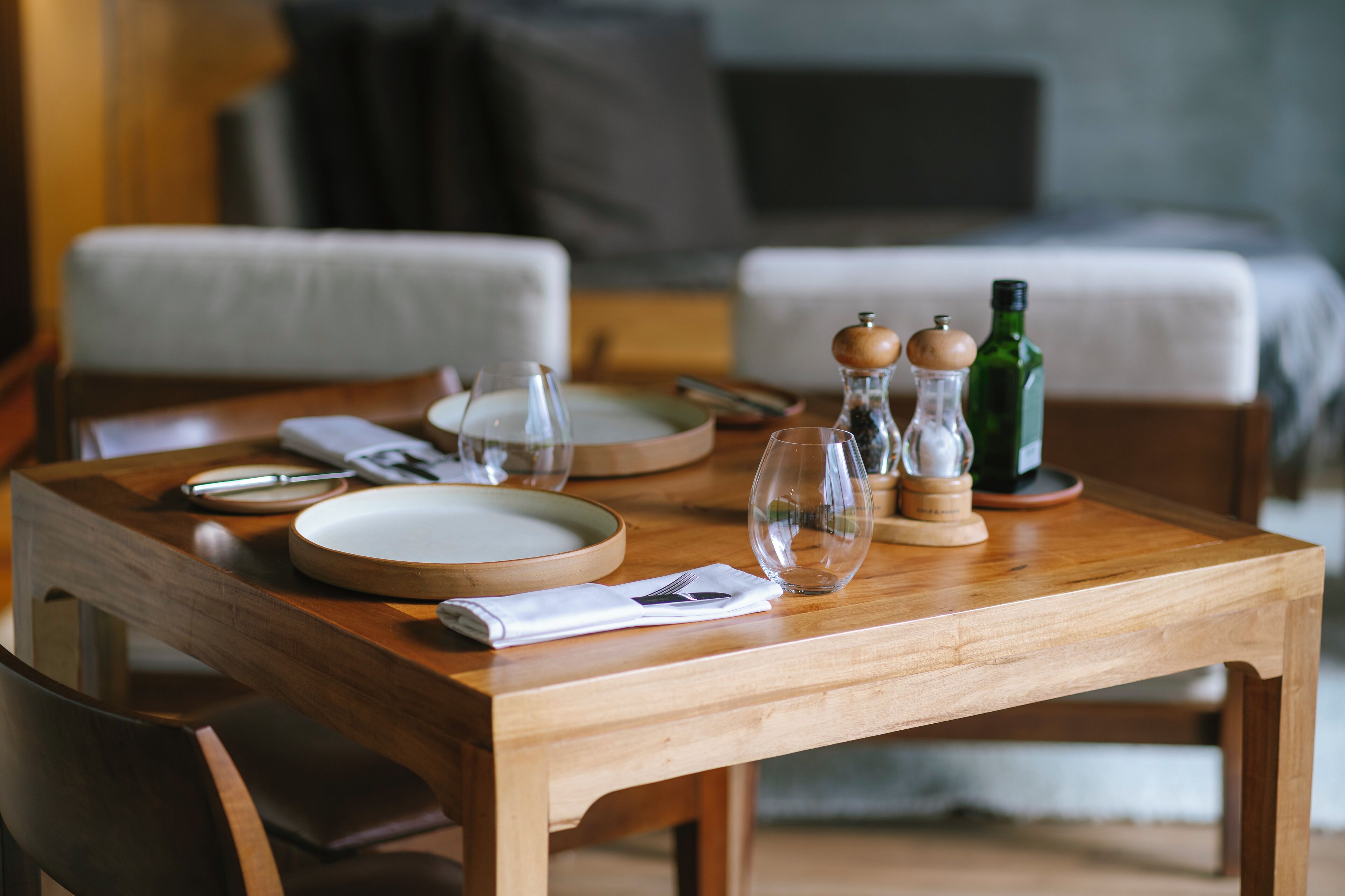 A wooden dining table set with plates, glasses, and a bottle, in a room with a bed and a chair.