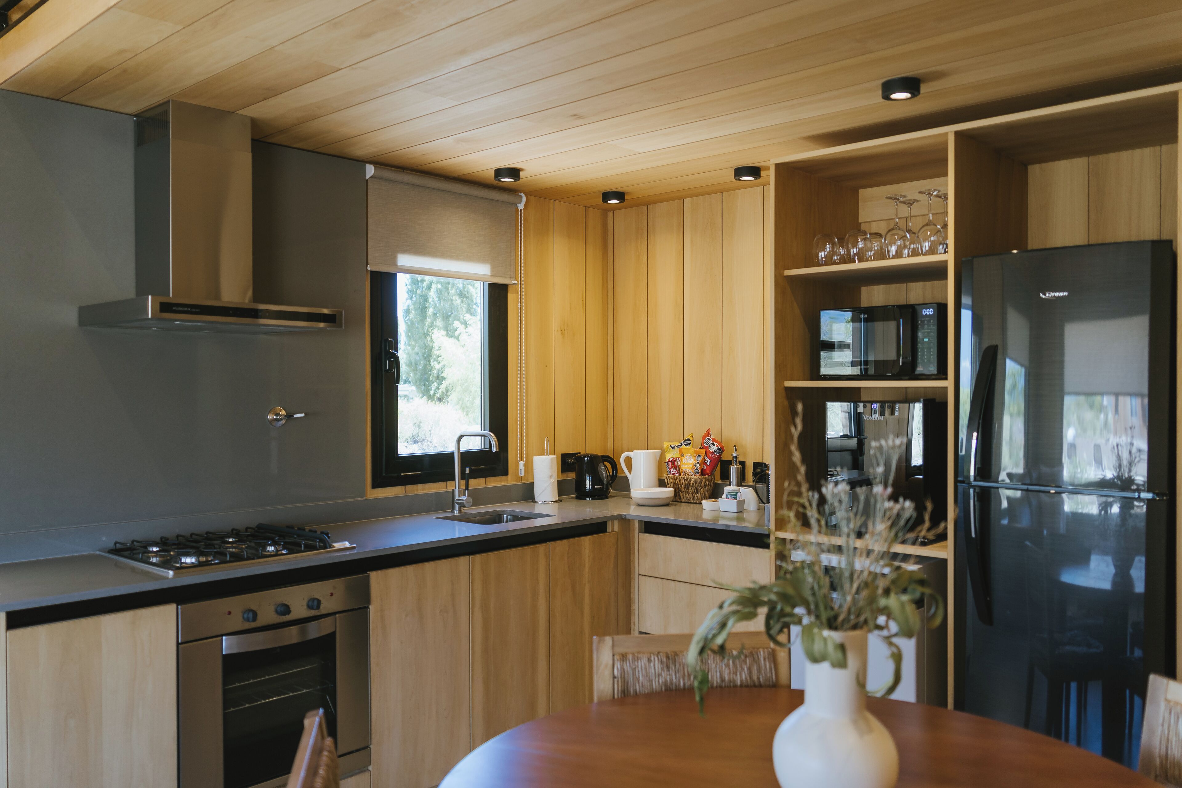 A modern kitchen with wooden cabinets, stainless steel appliances, and a dining area with a vase of dried flowers.