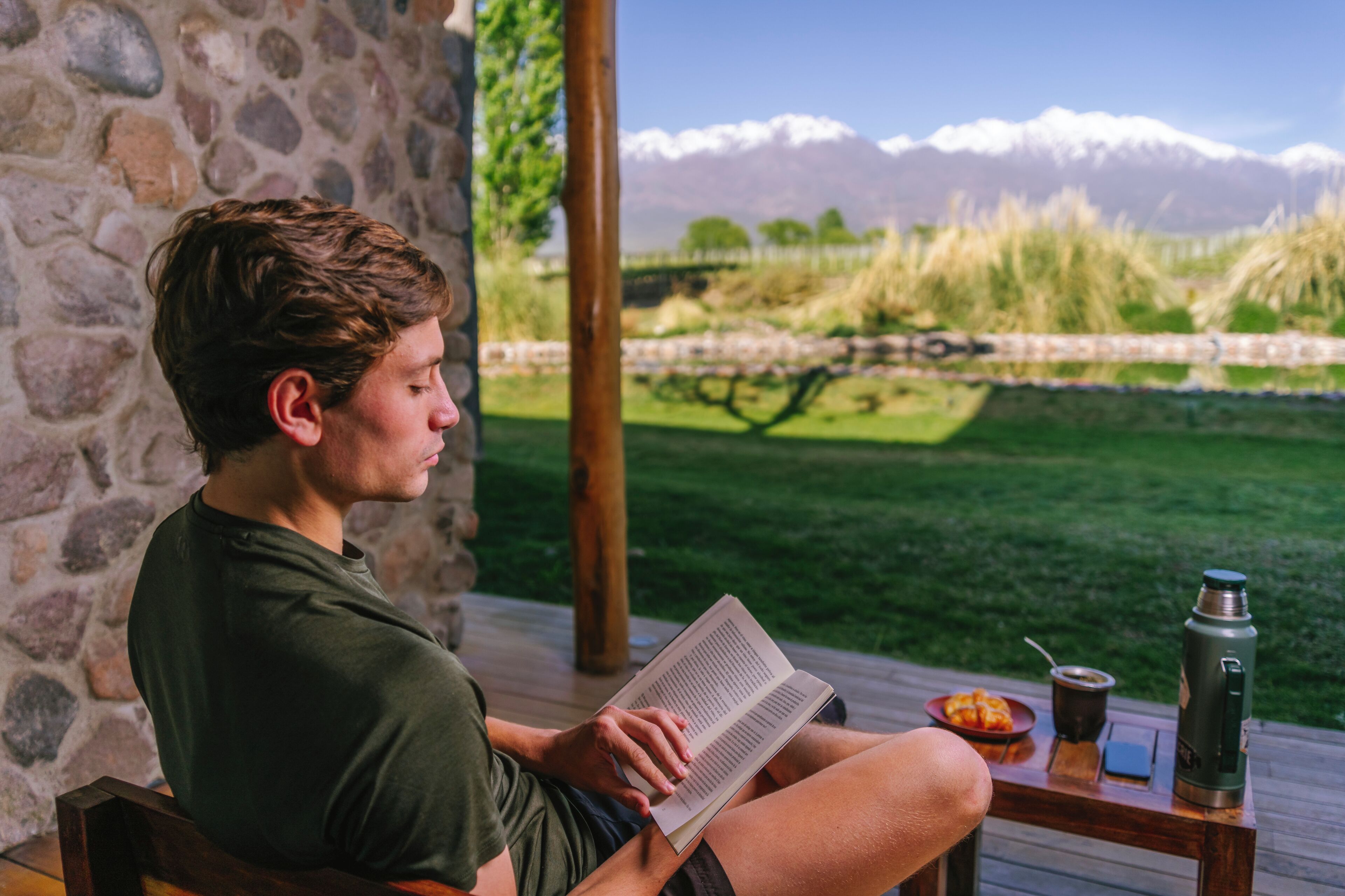 Una persona legge un libro su un pontile di legno con un muro in pietra, un tavolo con un thermos, un piatto di cibo e una vista sulle montagne e sull'erba.
