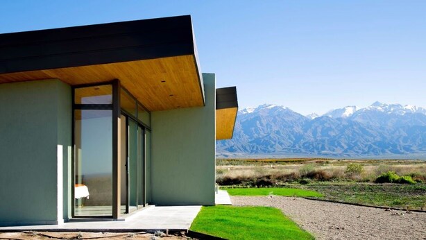 A modern building with a large glass door, a wooden overhang, and a view of mountains in the background. - La Morada Lodge - Valle de Uco (Tunuyan)