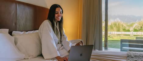 A person sitting on a bed with a laptop, in a room with a large window and a view of a landscape.