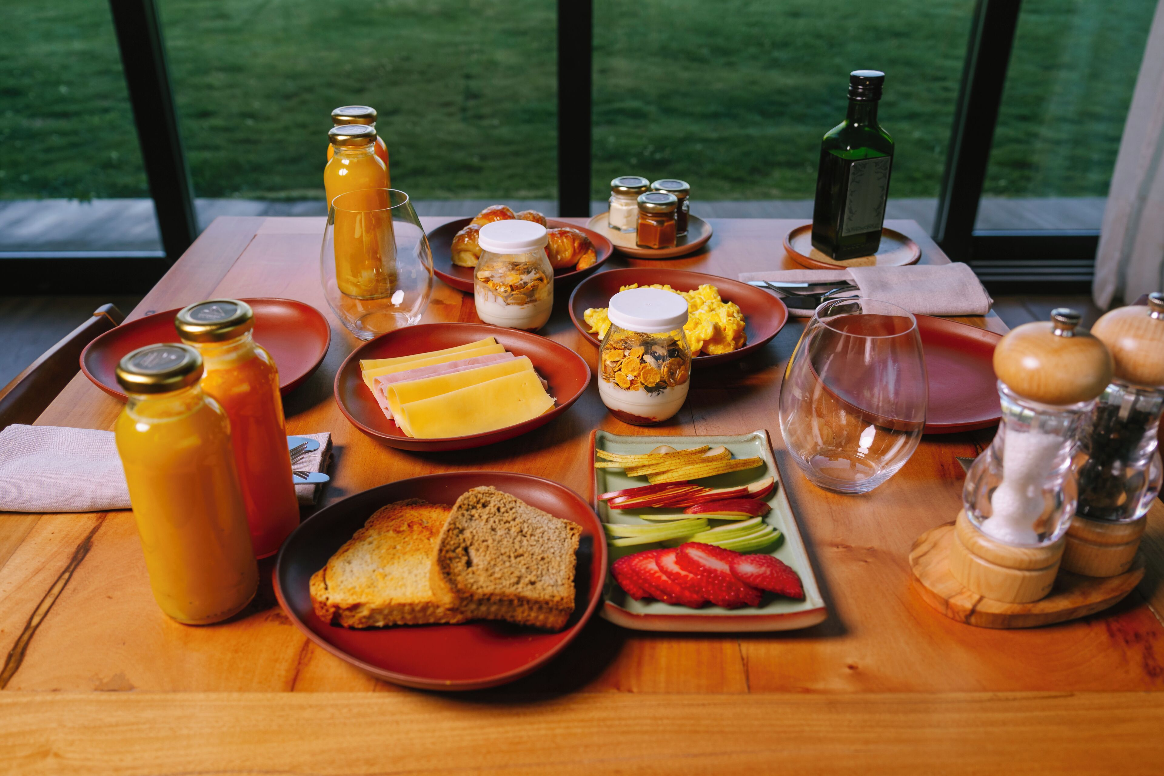A breakfast table with various food items and condiments, set on a wooden table with a view of a green landscape through large windows.