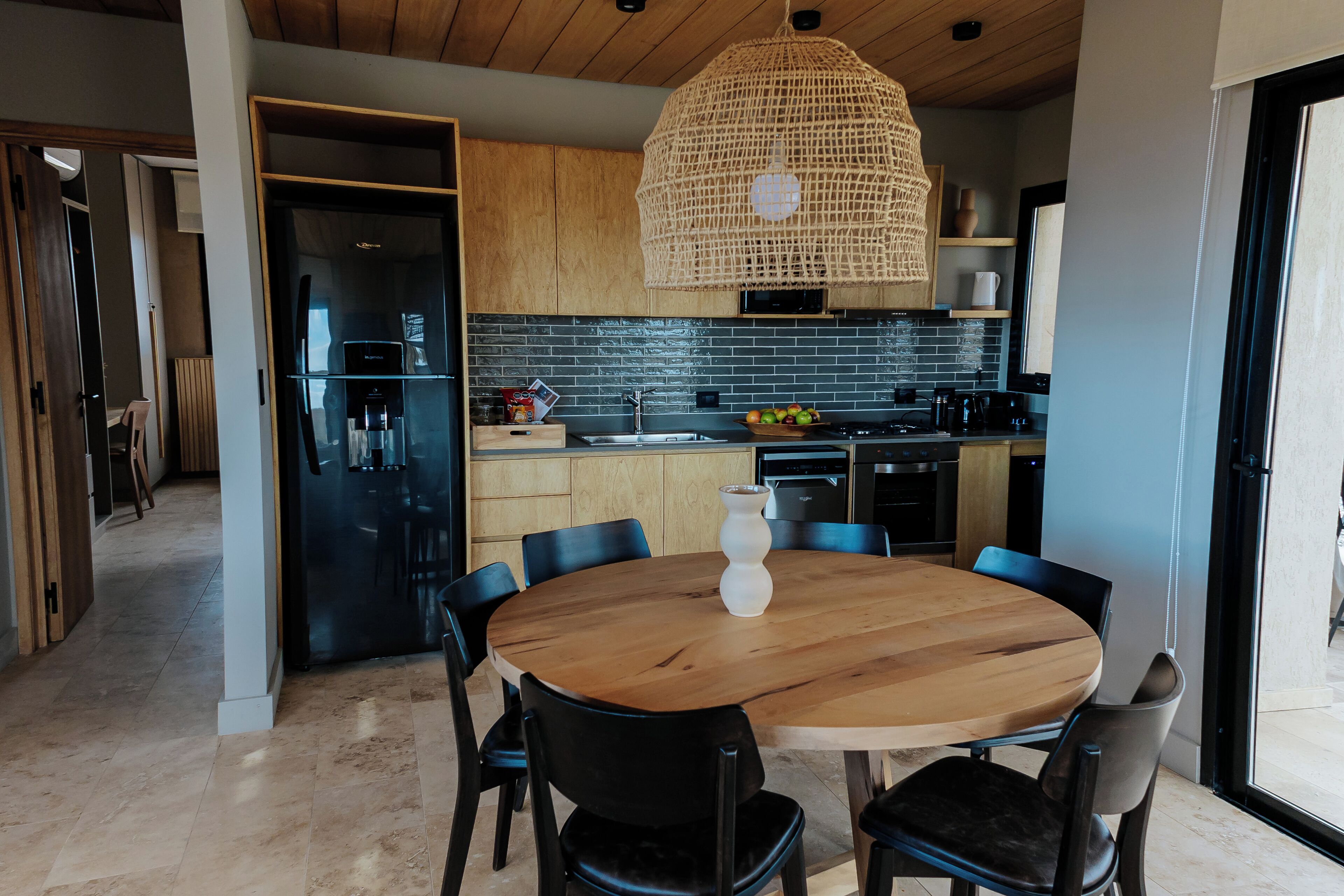 A modern kitchen with a round wooden table, black chairs, and a woven pendant light.