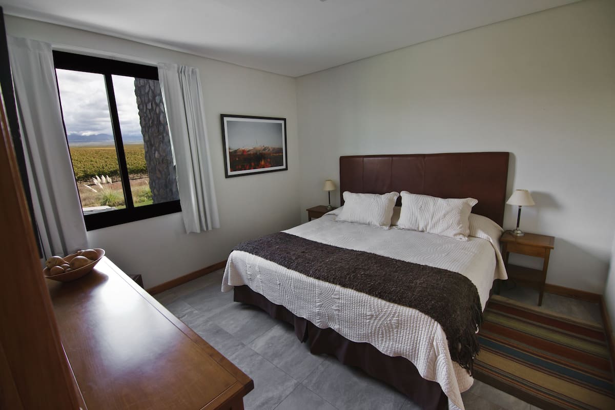A bedroom with a large bed, a nightstand, a lamp, a window with curtains, and a framed picture on the wall.