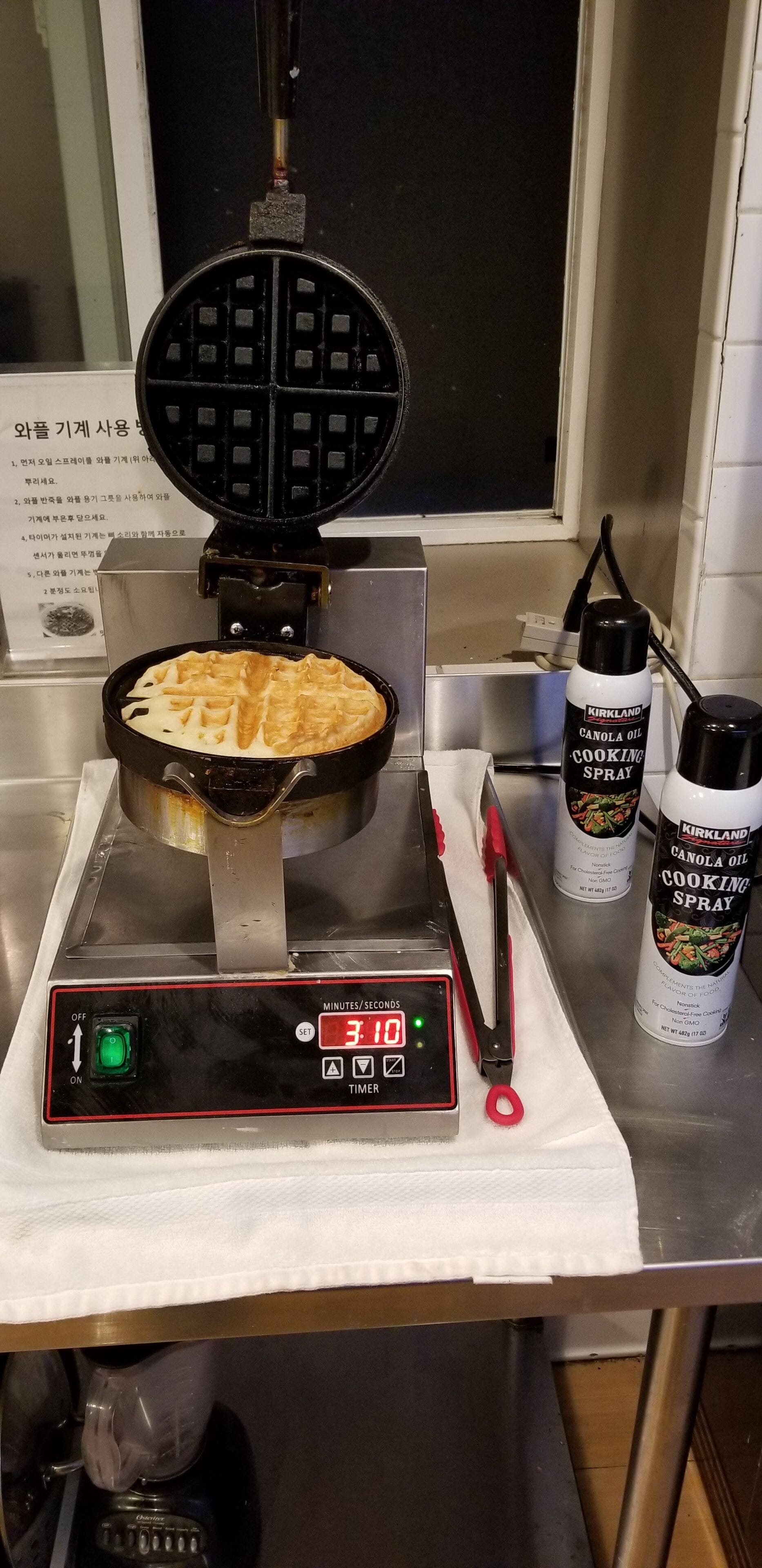 Un appareil à gaufres contenant une gaufre à l’intérieur, une minuterie et deux vaporisateurs d’huile de canola Kirkland sur un plan de travail de cuisine.