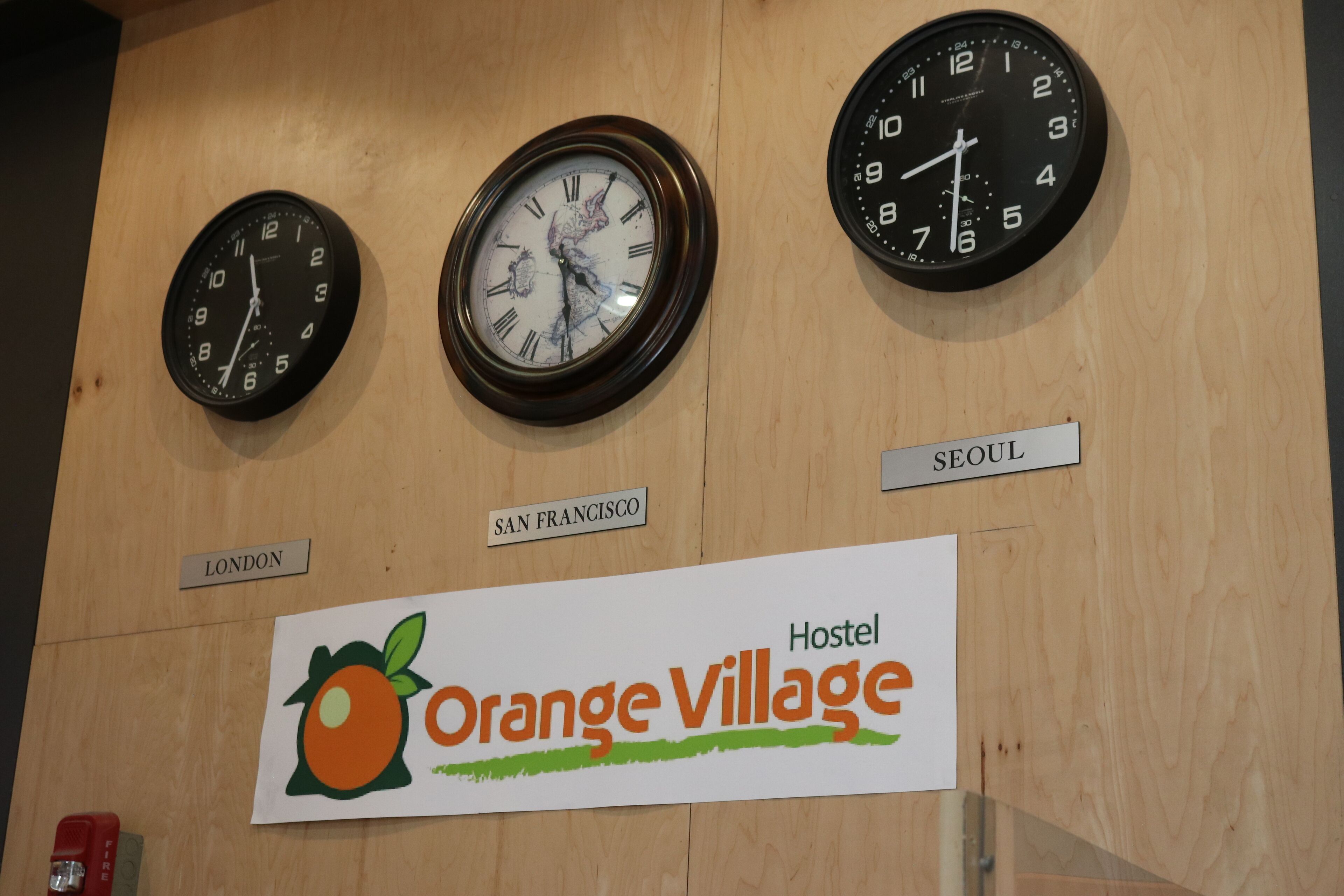 Trois horloges indiquant l’heure de Londres, de San Francisco et de Séoul sont accrochées à un mur en bois. En dessous, une pancarte indique : « Auberge Orange Village ».