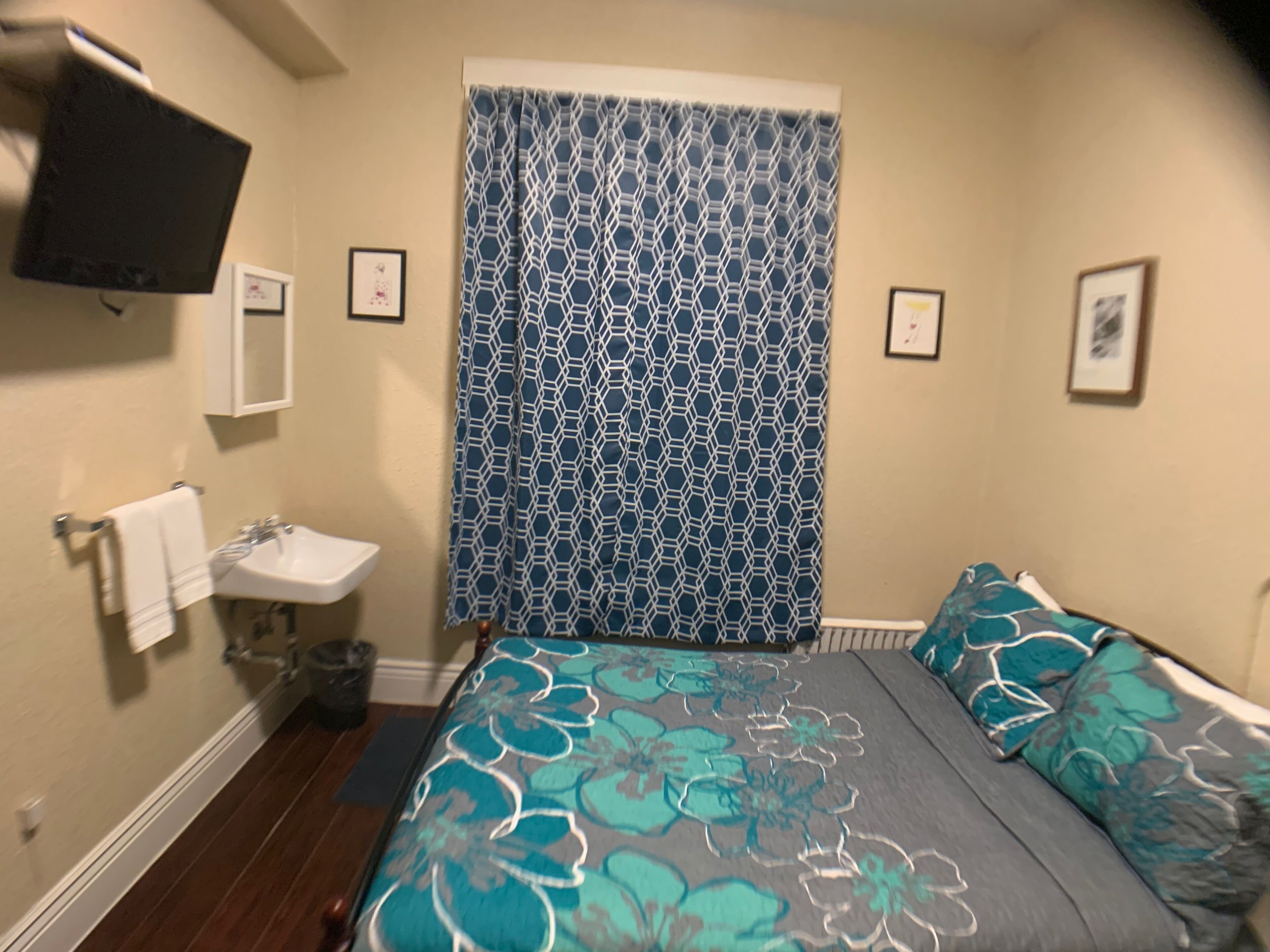 A bedroom with a bed, a bedside table, a wall-mounted TV, a sink, and a window with a patterned curtain.