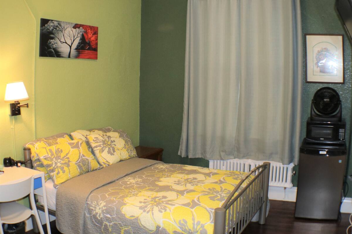 A bedroom with a bed, a chair, a table, a lamp, a radiator, and a microwave.