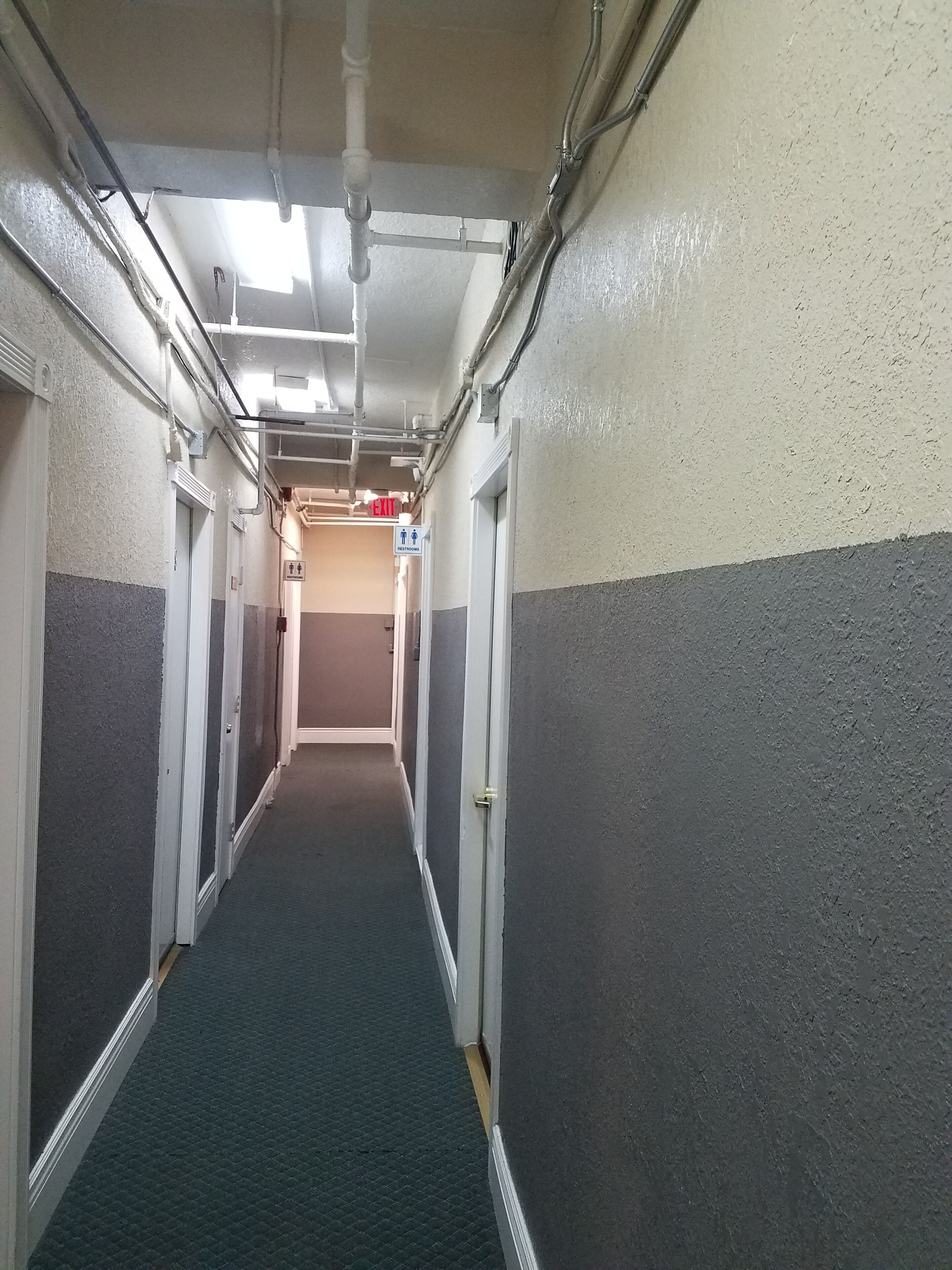 A narrow hallway with a carpeted floor, walls painted in two-tone, and an exit sign visible at the end.