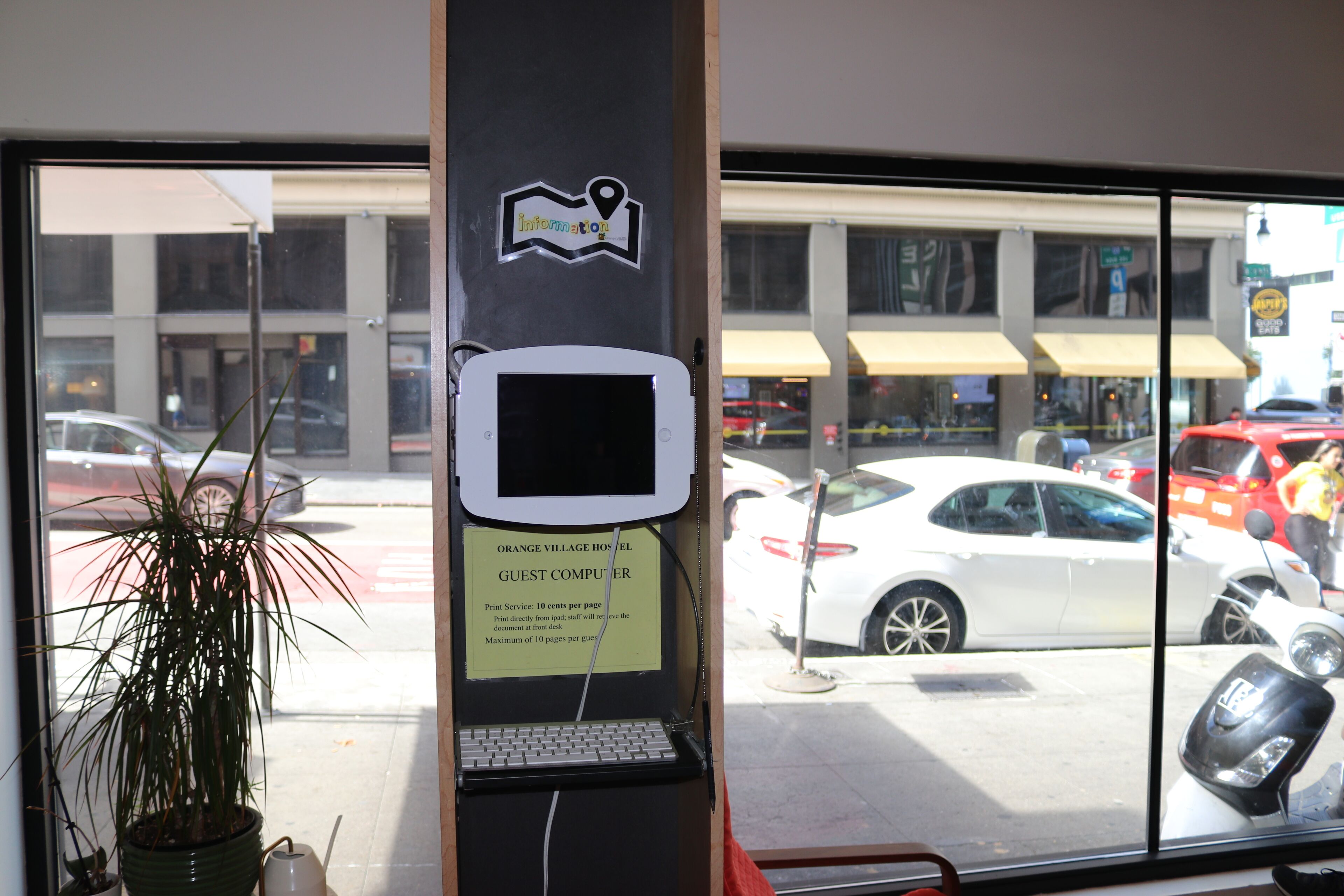 A guest computer kiosk with a location pin icon, a sign for Orange Village Hotel, and a view of parked cars outside.