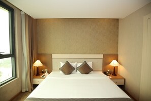 A hotel room with a large bed, two bedside tables with lamps, a headboard, and a window with curtains.