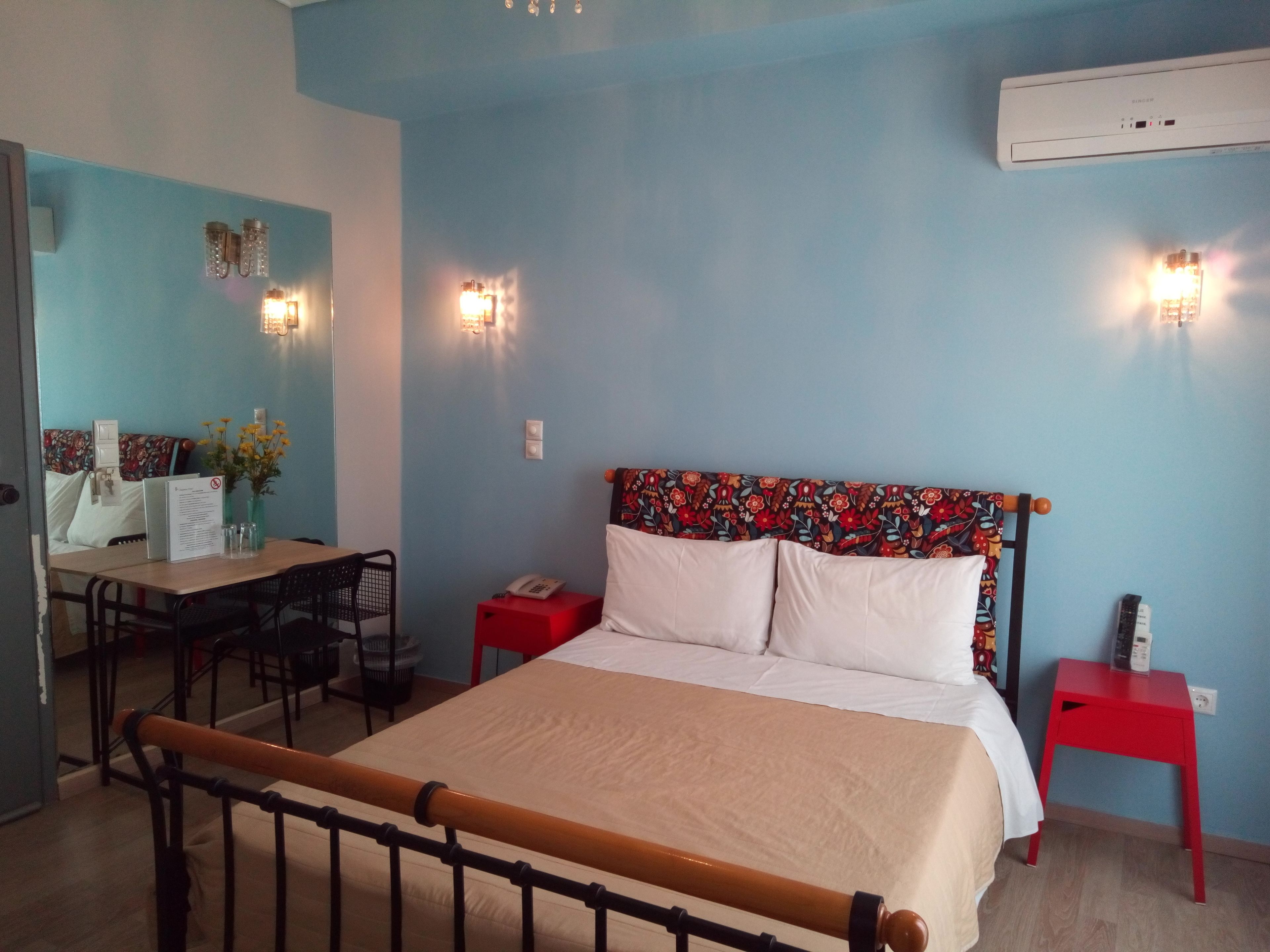 Une chambre d’hôtel avec un lit, un bureau et une chaise. On y trouve une table de chevet rouge avec un téléphone et une petite table d’appoint rouge avec une lampe.
