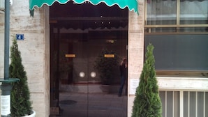 Entrada de hotel con toldo verde, dos árboles siempreverdes en macetas y un felpudo con la palabra 'Welcome'.