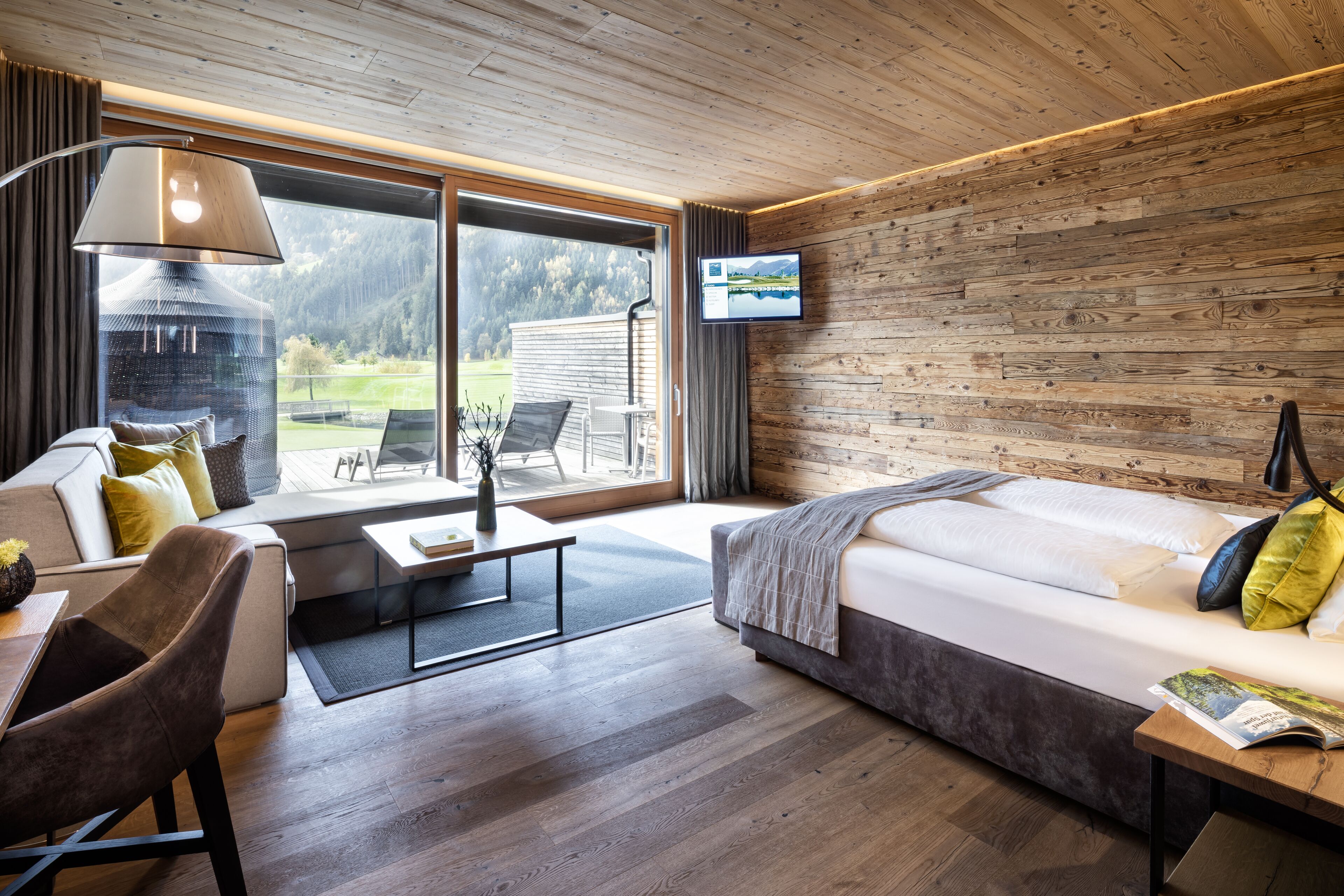 A modern room with a large bed, a sofa, a TV mounted on the wall, and a view of the outdoors.
