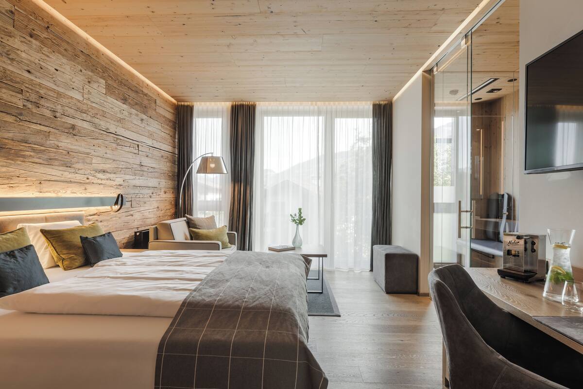 A modern hotel room with a wooden ceiling, a large bed, a desk with a coffee maker, and a TV mounted on the wall.