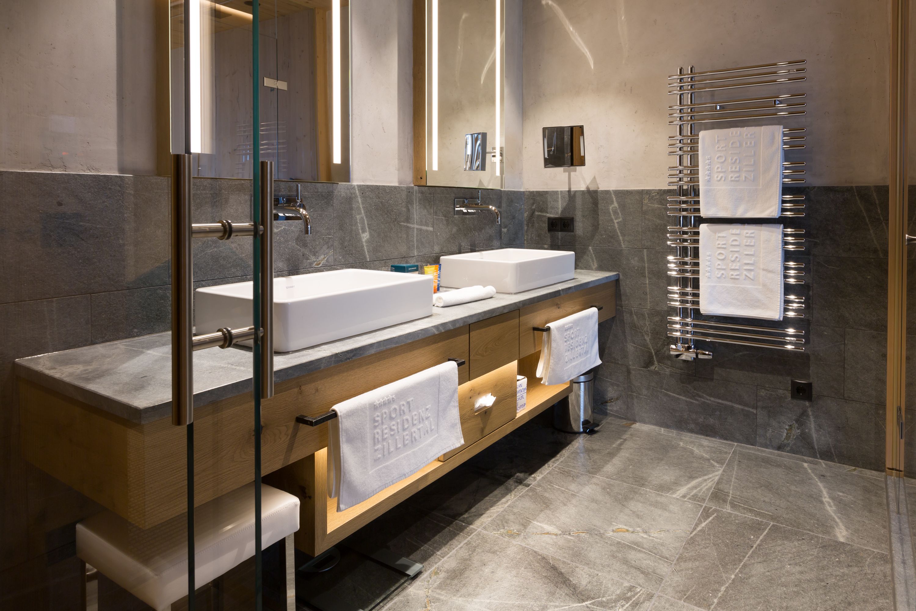Un baño moderno con dos lavamanos, cabina de ducha de vidrio y un perchero con varias toallas.