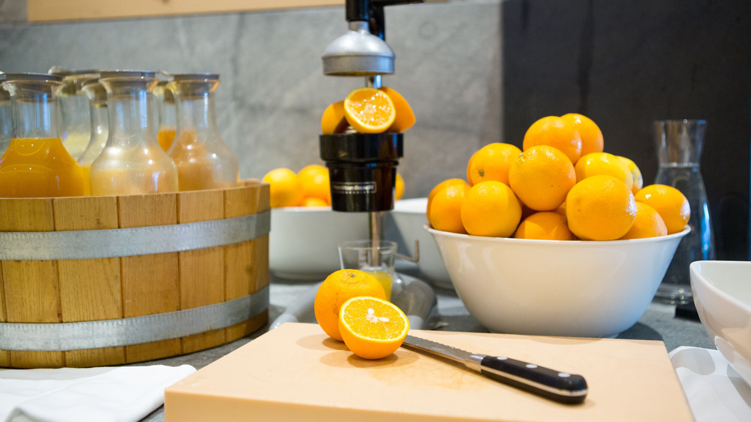 Una encimera de cocina con una cesta de madera que contiene botellas, un bol de naranjas y una tabla de cortar con un cuchillo.