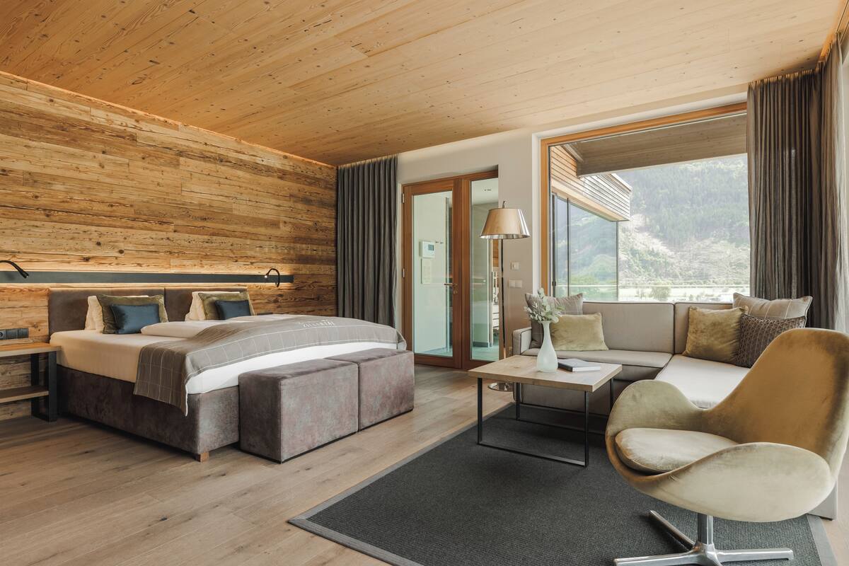 A modern hotel room with a wooden ceiling, a large bed with a headboard, a sofa, a small table, and a view of the outdoors.