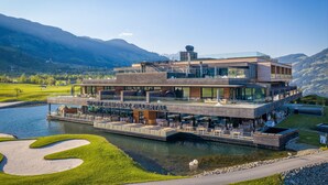 Un hôtel moderne doté d’une façade en verre, d’une terrasse et offrant une vue sur un lac et des montagnes.