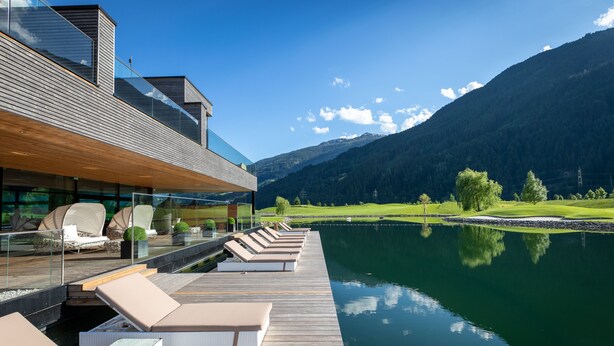 Un bâtiment moderne doté d’une terrasse en bois qui offre une vue sur un lac paisible, le tout étant entouré de montagnes.
