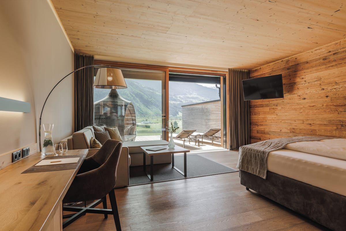 A modern hotel room with a wooden ceiling, a large window offering a mountain view, a bed, a desk with a lamp, and a TV mounted on the wall.