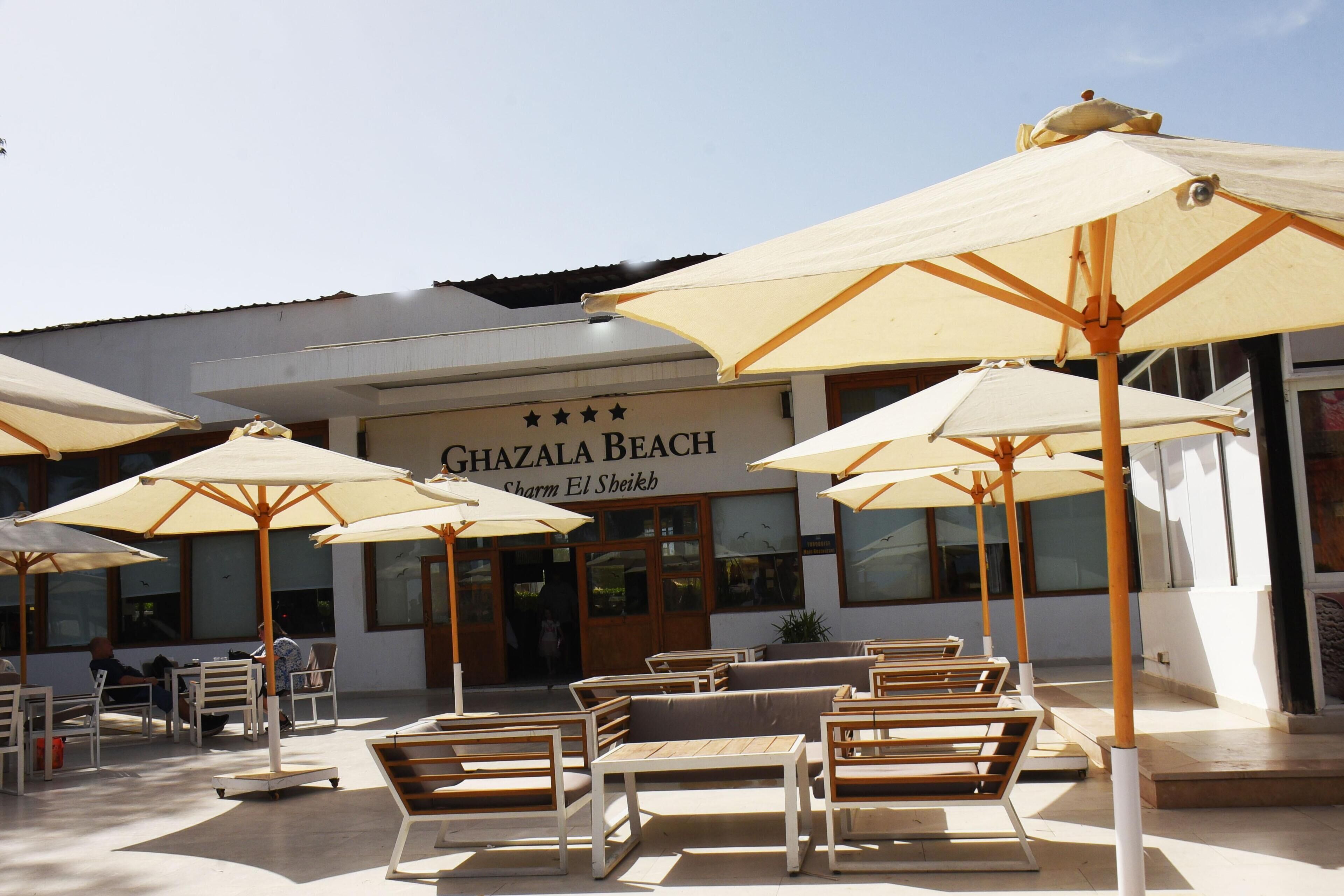Outdoor seating with umbrellas, a building with a sign reading 'GHAZALA BEACH,' and a clear sky.