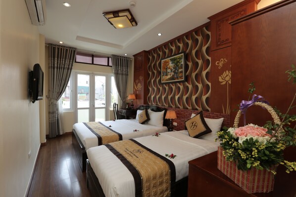 A hotel room with two beds, a TV, a window with curtains, a wall-mounted picture, and a decorative basket with flowers.