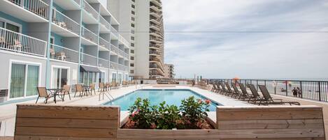 A poolside area with lounge chairs, a wooden planter with flowers, and a tall building in the background.