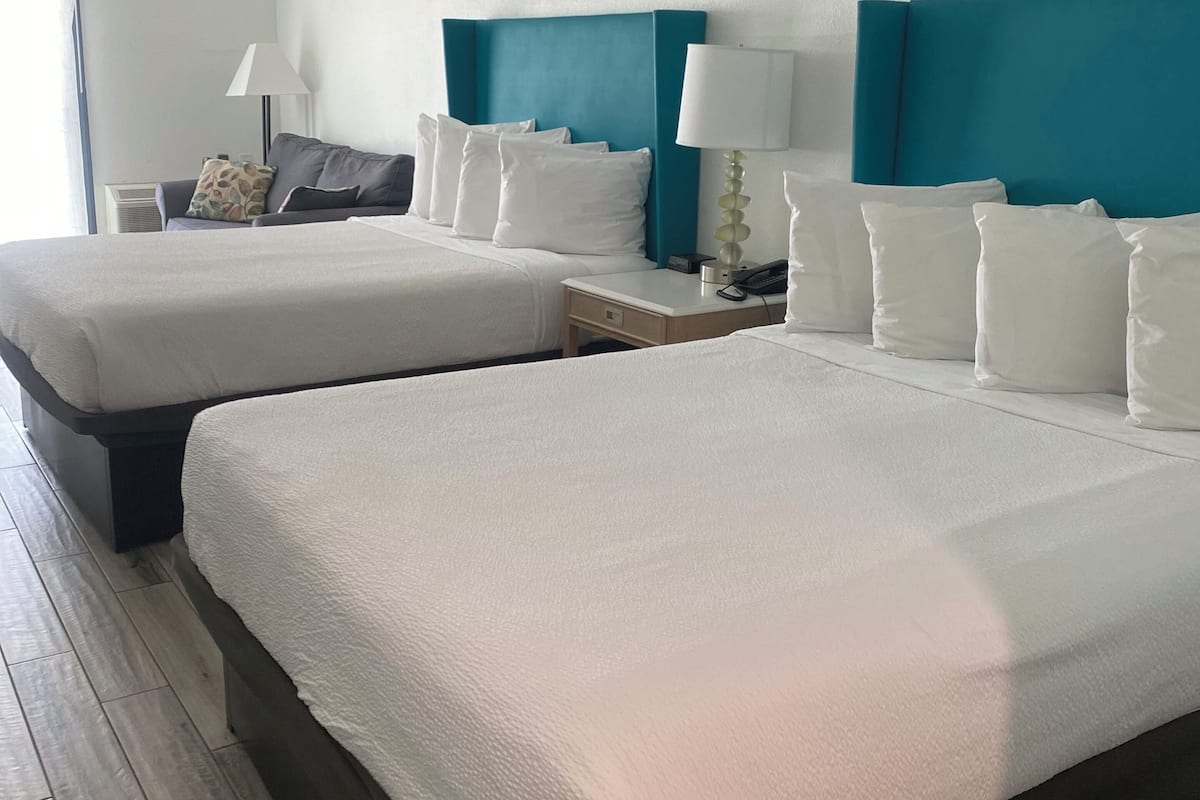 A hotel room with two beds, a nightstand with a lamp, a sofa, and a window with curtains.