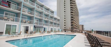 A swimming pool with lounge chairs and tables, a multi-story building with balconies, and a clear sky.