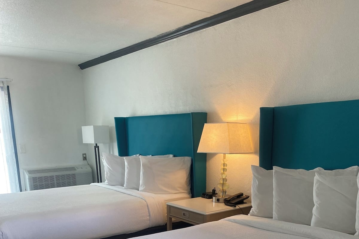 A hotel room with two beds, a nightstand, a lamp, and an air conditioning unit.