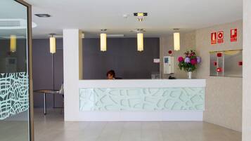 Un área de recepción con diseño moderno, que incluye un mostrador de recepción, un detector de incendios instalado en la pared y un arreglo floral decorativo.