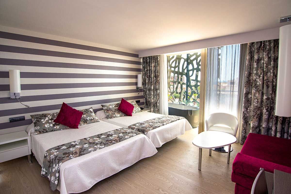 A hotel room with two beds, a chair, and a view of the outdoors.