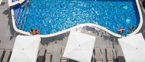 Una piscina con gente nadando y sillas de descanso junto con sombrillas.