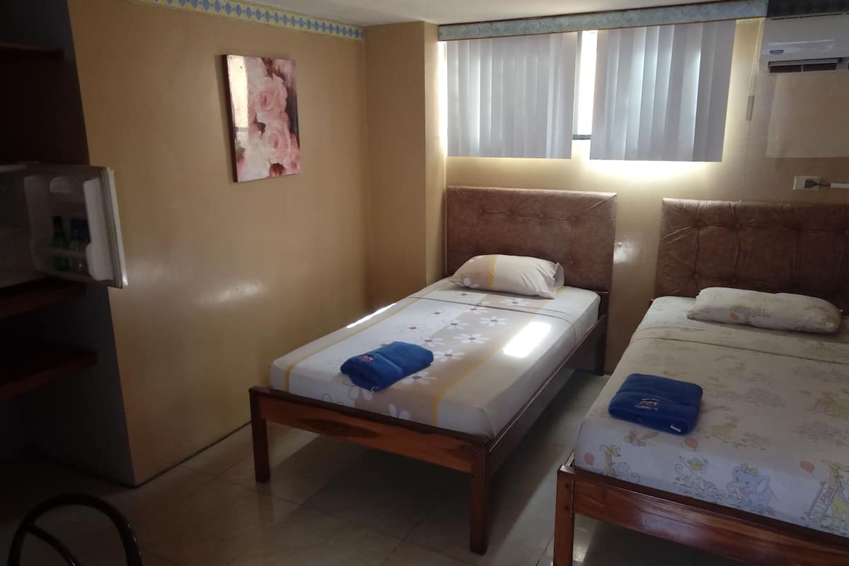 A room with two beds, a wall-mounted air conditioner, a window with blinds, and a small table with a bottle.