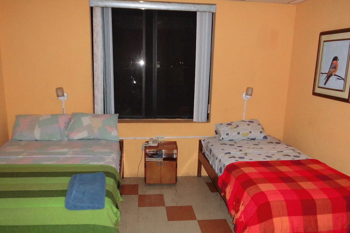 A room with two beds, a window, a nightstand, and a wall-mounted lamp.