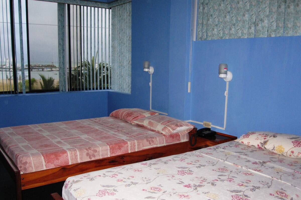 A room with two beds, a window with blinds, a telephone, and a wall-mounted light fixture.
