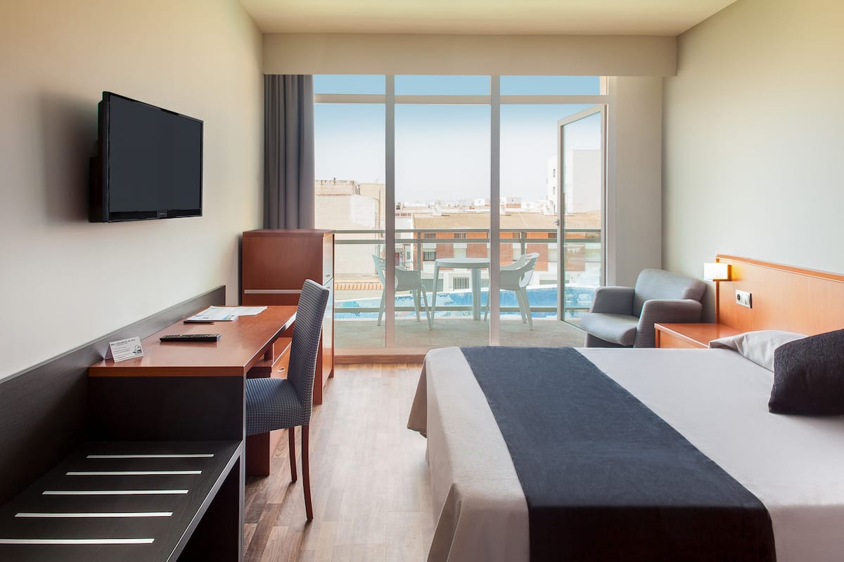Habitación de hotel con cama, escritorio, silla, televisor y balcón con vista a la piscina.
