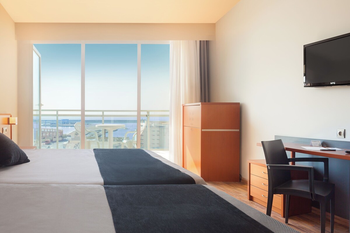 A hotel room with two beds, a desk, a chair, a TV, and a large window with a city view.