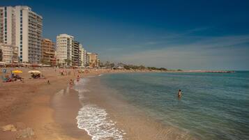Una playa con una hilera de edificios altos, cielos despejados y mar tranquilo.