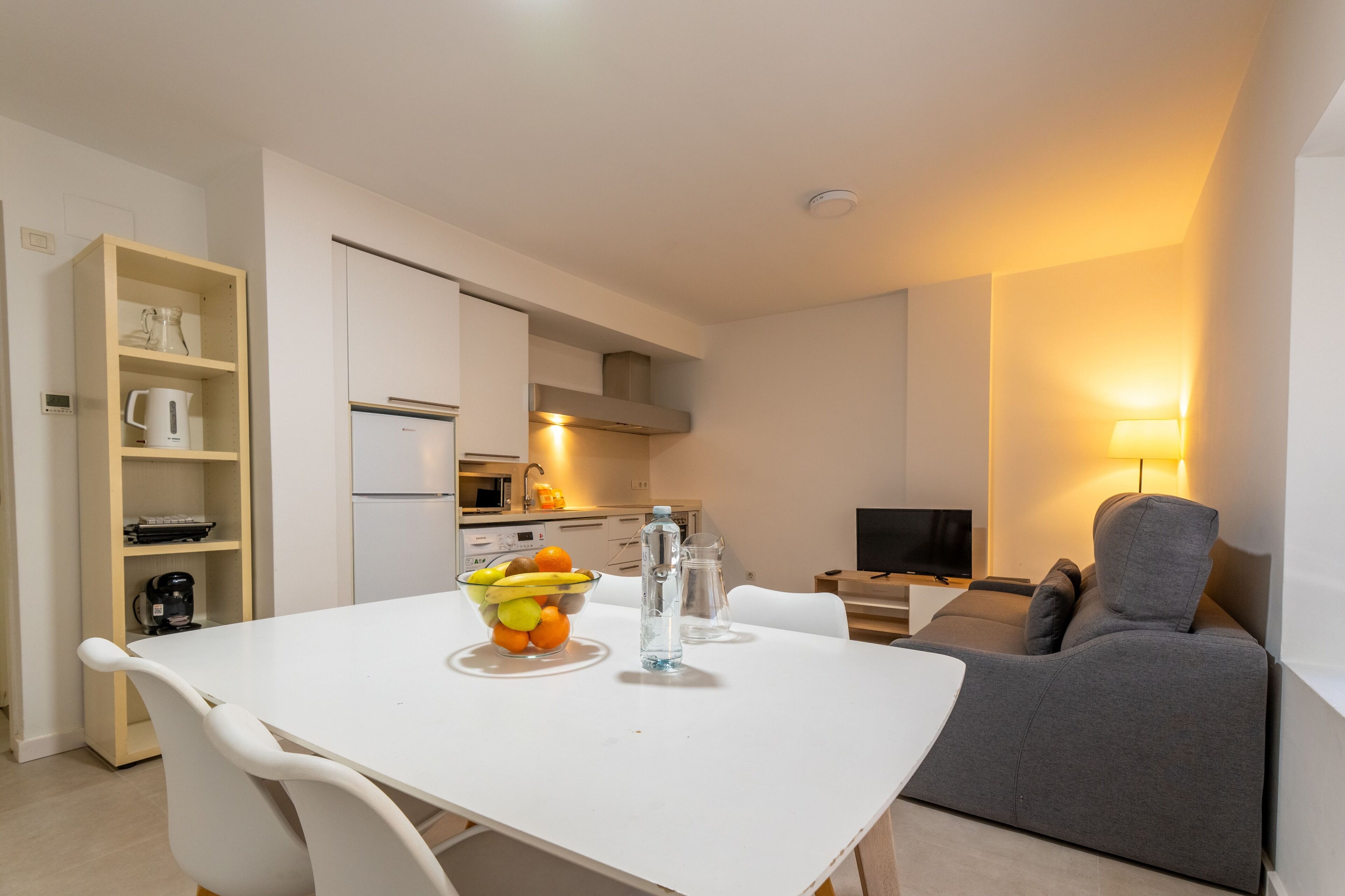 Een moderne keuken met witte kasten, een eettafel met stoelen, een schaal fruit en een waterfles.