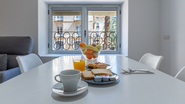 A dining table with breakfast items, a window with a view of buildings, and a balcony railing. - Apartamentos Santa Faz by Be Alicante (Alicante)
